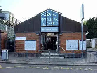 A brown-bricked building with a rectangular, light blue sign reading "WEST HARROW STATION" in white letters all under a light blue sky