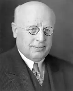 A photographic portrait of a balding white man with small round glasses